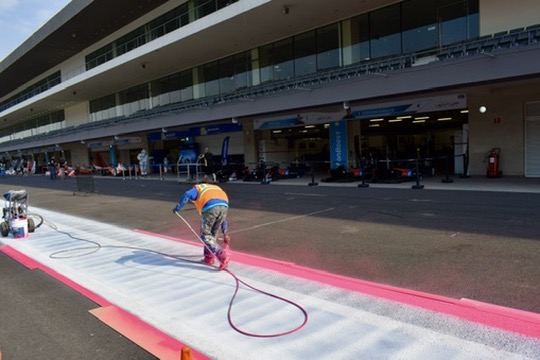 A ground crew member using a professional spray machine to apply a temporary, vibrant pink line to the asphalt of a Formula E street circuit, with blurred Gen3 electric race cars in the background.