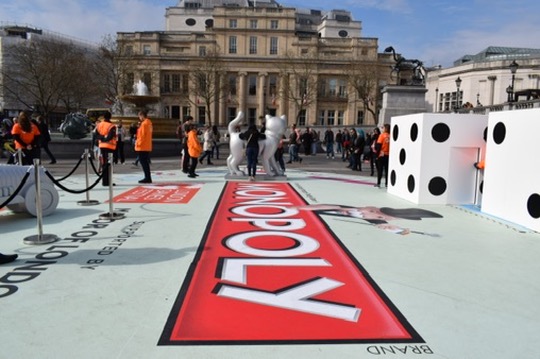 A wide-angle view of the giant Monopoly board at Trafalgar Square, showing people interacting with life-size playing pieces and the custom-printed floor graphic.