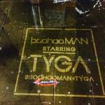 A clean graffiti pavement stencil on wet, dark stone at night. The stenciled text, enclosed in a square border, reads "boohooMAN STARRING TYGA" followed by the hashtag "#BOOHOOMANxTYGA". Reflections from streetlights highlight the cleaned lettering.