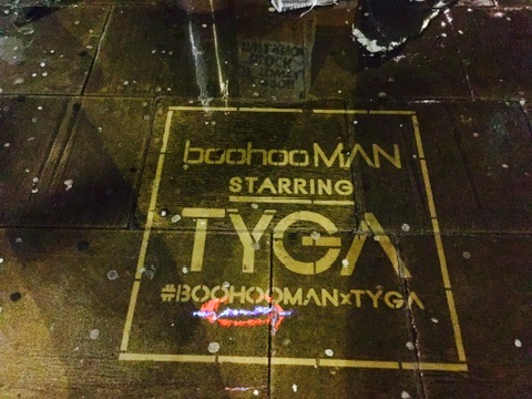 A clean graffiti pavement stencil on wet, dark stone at night. The stenciled text, enclosed in a square border, reads "boohooMAN STARRING TYGA" followed by the hashtag "#BOOHOOMANxTYGA". Reflections from streetlights highlight the cleaned lettering.