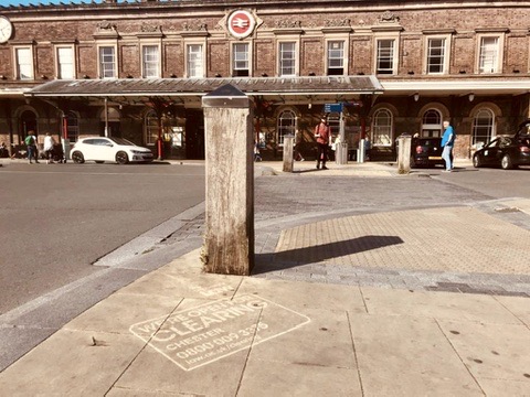 A clean graffiti stencil advertisement on the pavement outside Chester train station. The white stencil text reads "We’re Open for Clearing, Chester, 0800 009 3376, law.ac.uk/clearing" for the University of Law. The historic station building is visible in the background under bright sunlight.
