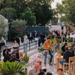 A bustling outdoor scene at the Frieze LA art fair, featuring attendees walking and socializing on black and white temporary painted stripes, surrounded by lush vendor greenery and event structures.