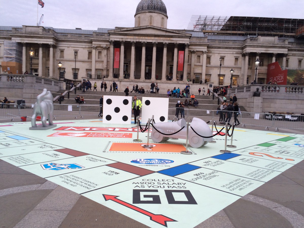 A giant temporary Monopoly board floor graphic installed on the paving at Trafalgar Square, London, featuring life-size, custom-built playing pieces including a car, dog, and large dice.