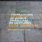 White and orange temporary eco-paint stencil on stone paving at Liverpool One, featuring a Tilda Swinton quote for a Tate Liverpool experiential marketing campaign.